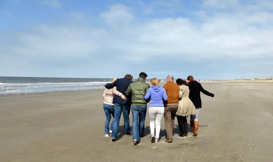 Een groep loopt arm in arm over het strand, van achteren gezien